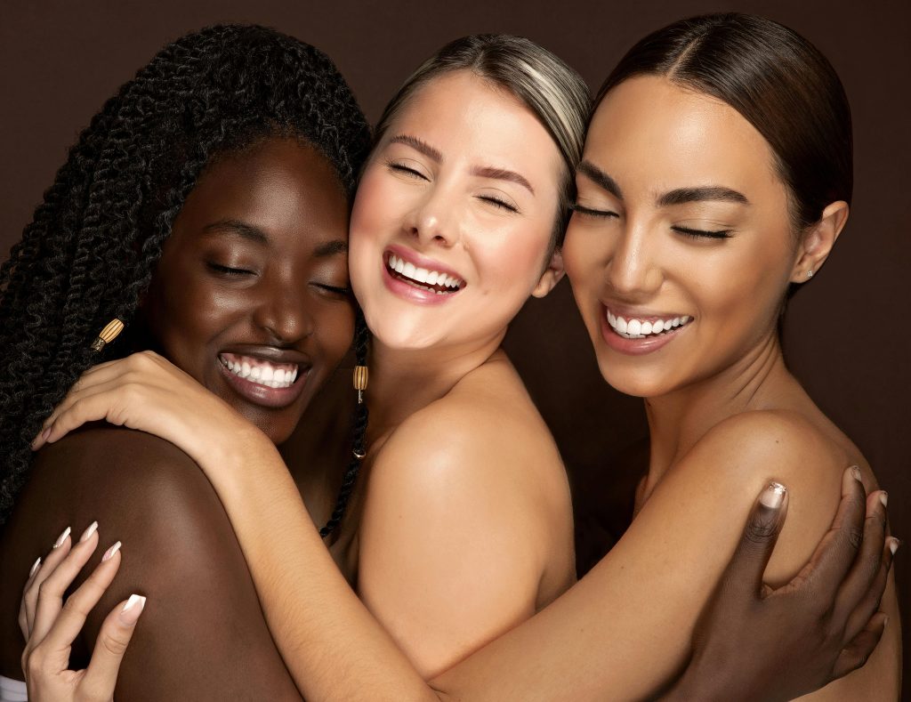 Three diverse women embracing with eyes closed and smiling, celebrating friendship and unity.